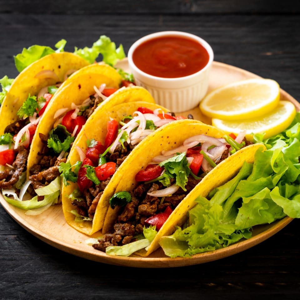 Five tacos with meat, lettuce, tomatoes, and onions on a wooden plate with salsa and lemon slices.